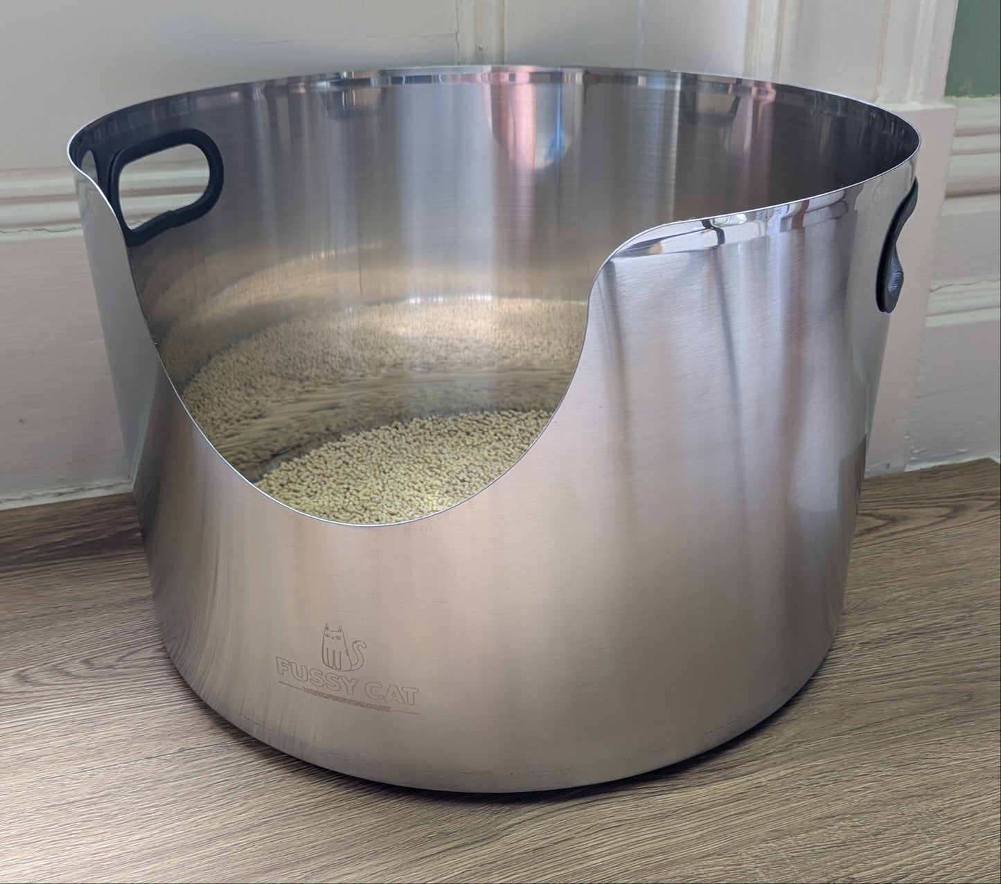 Stainless steel round cat litter box on a wooden floor with a white wall background