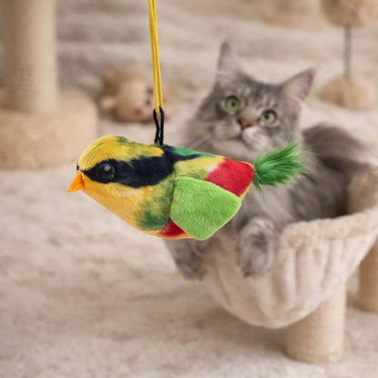 Plush yellow, green, and red chirping bird cat toy with a green tail, hanging by a yellow string, with a grey cat in the background.