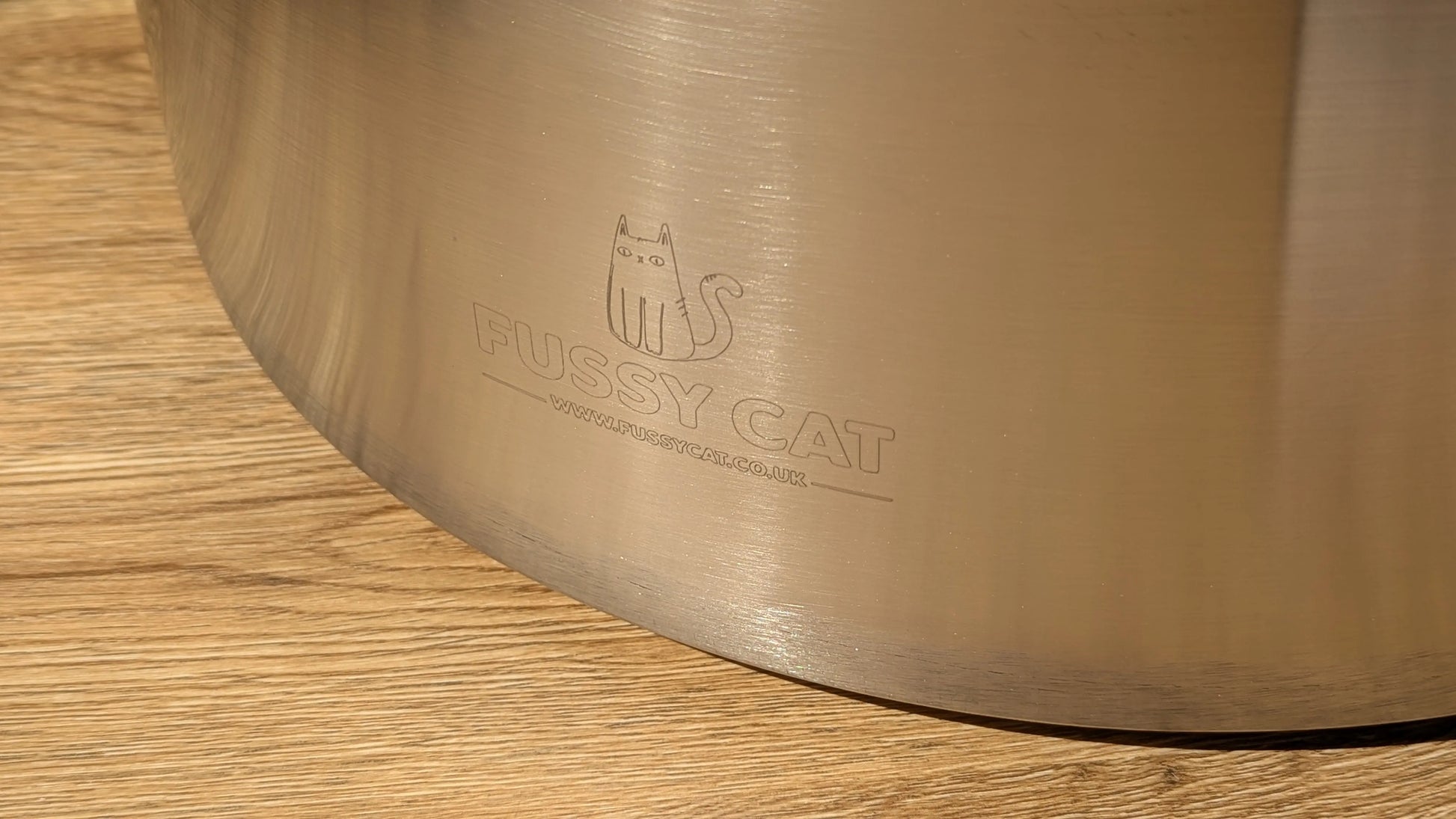 Close-up of the Fussy Cat round stainless steel litter box with engraved logo on a wooden floor.
