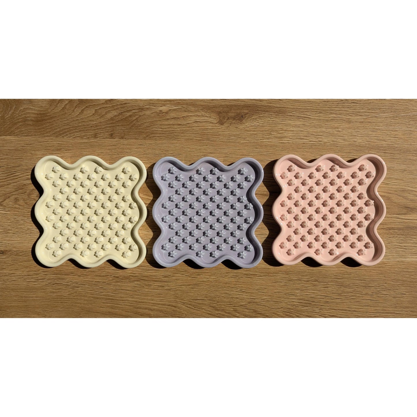 Three ceramic Fussy Cat slow feeders in pastel yellow, purple, and pink, with wavy edges and flower-shaped nubs.