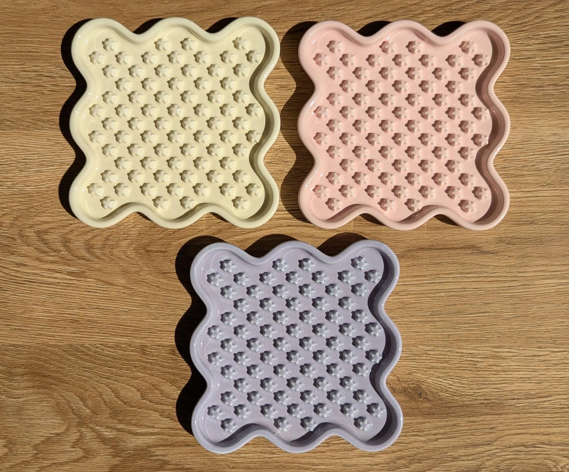 Three ceramic Fussy Cat slow feeders in pastel yellow, pink, and purple, featuring a wavy square shape and flower-like food compartments.