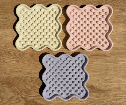 Three ceramic Fussy Cat slow feeders in pastel yellow, pink, and purple, featuring a wavy square shape and flower-like food compartments.