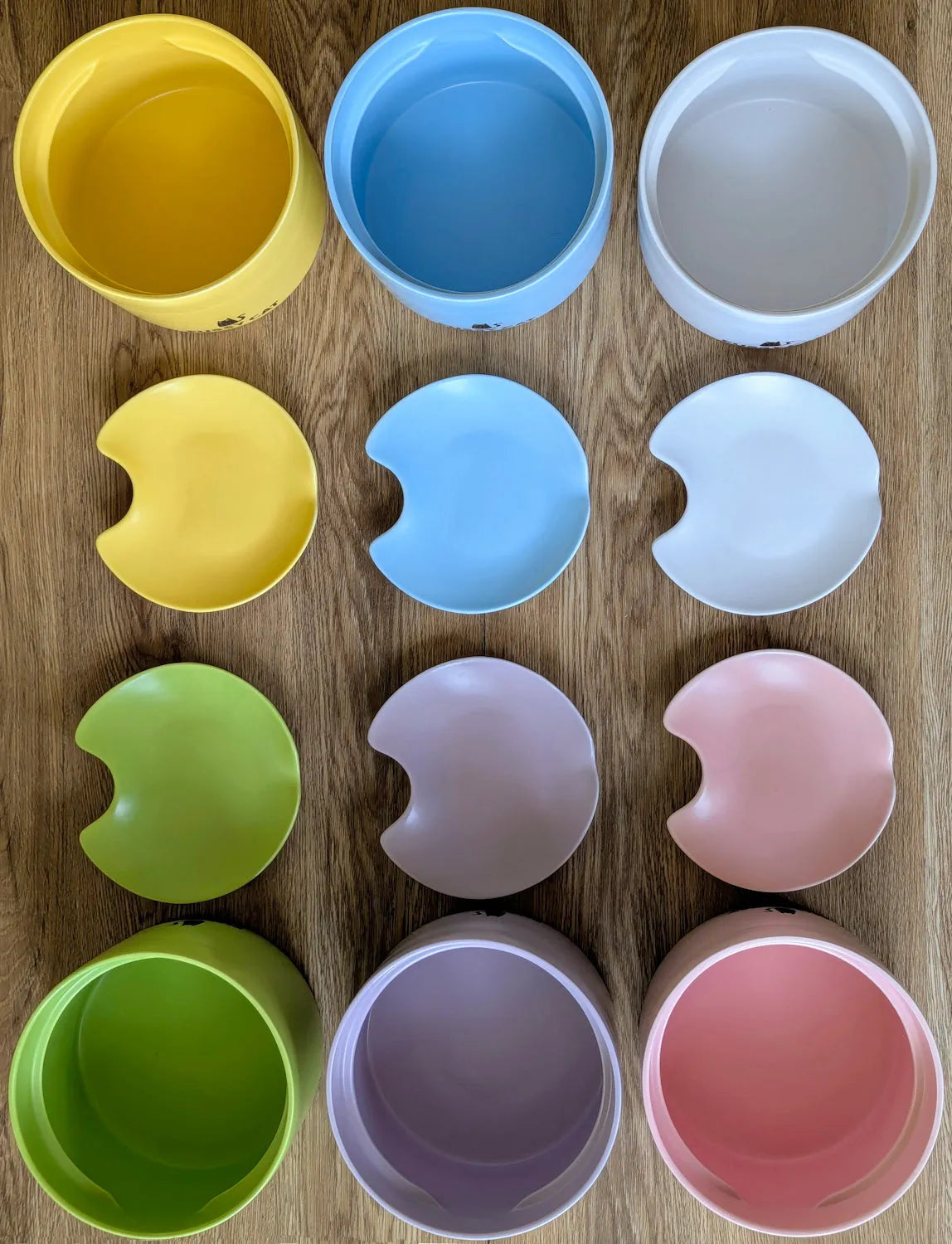 Six Fussy Cat ceramic water fountain bases and six matching spill-resistant lids in pastel yellow, blue, white, green, lavender, and pink, arranged on a wooden surface.