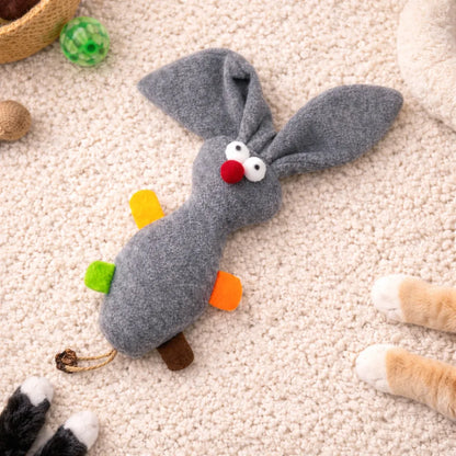 Grey plush rabbit cat toy with red nose, white eyes, colorful felt accents, and a braided tail on a cream rug.