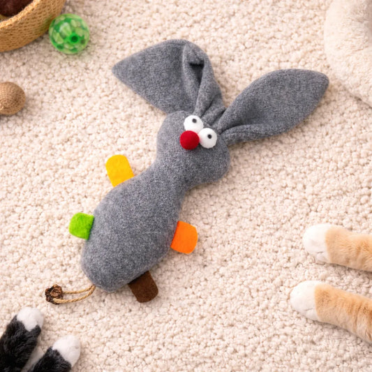 Grey plush rabbit cat toy with red nose, white eyes, colorful felt accents, and a braided tail on a cream rug.