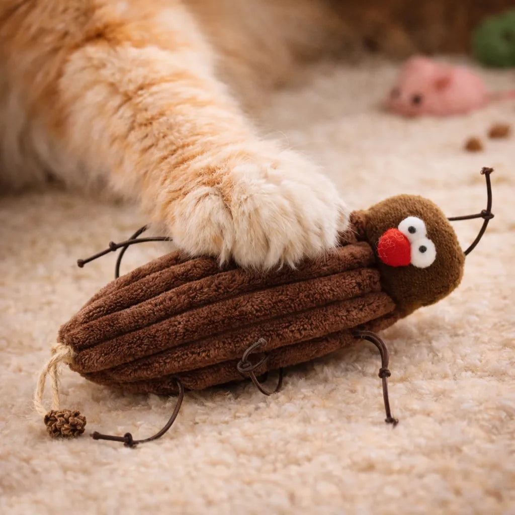 Brown plush cockroach cat toy with red nose, white eyes, and wire legs, held by a cat's paw on a beige rug.